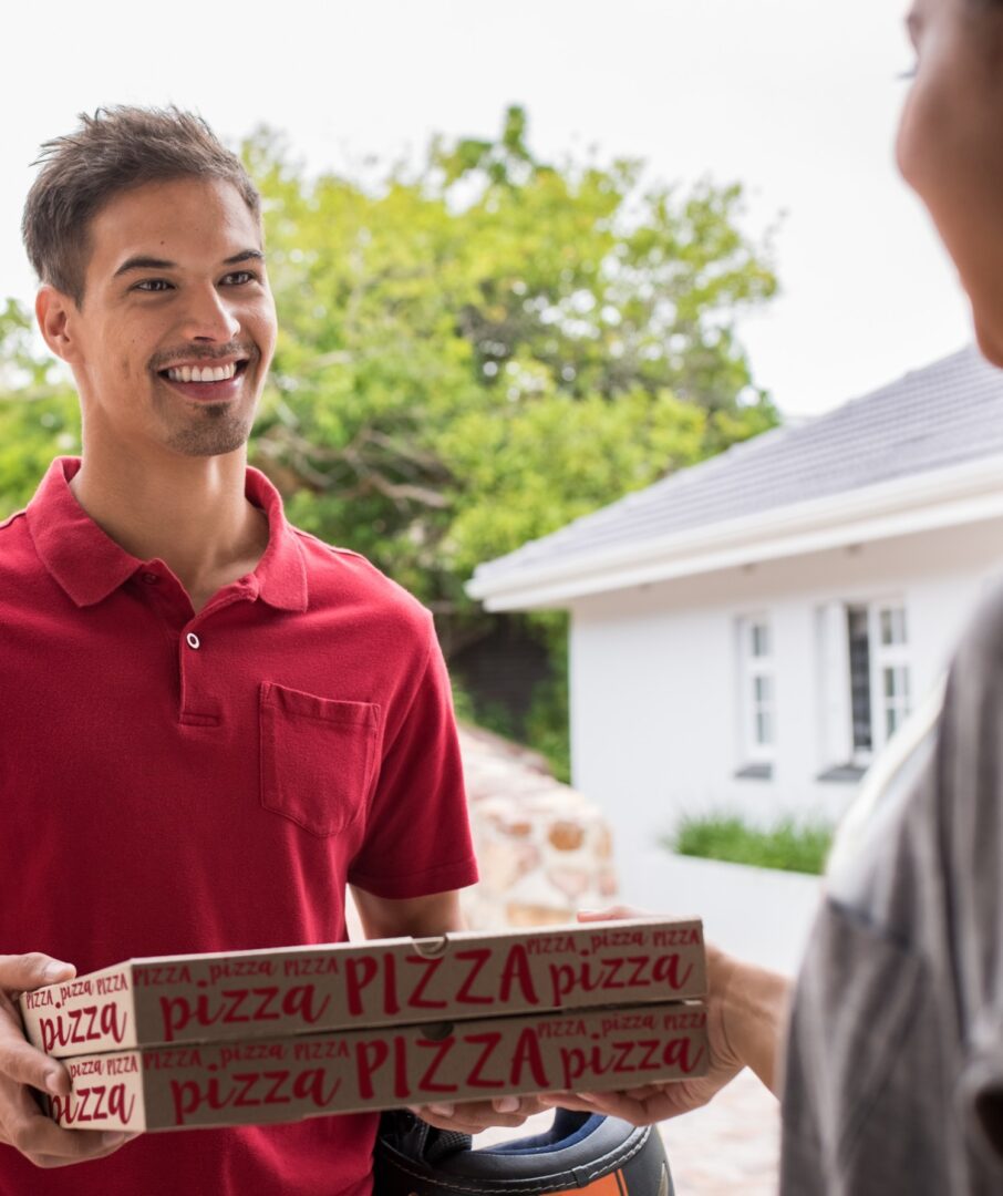 A smiling person in a red shirt holding pizza boxes at a doorstep, engaging with another person. House and trees are visible in the background.